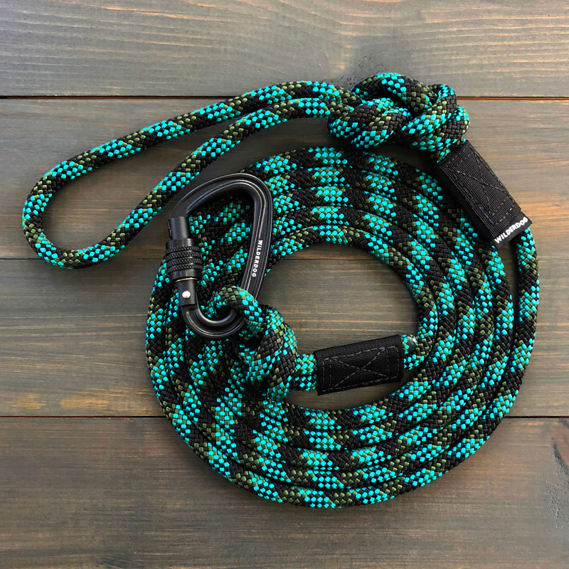 A coiled black, green, and turquoise Granite Leash lies on a wooden surface. The leash is fitted with a black metal carabiner clip and features reinforced black fabric accents at various points for durability, making it perfect for adventurous outings.